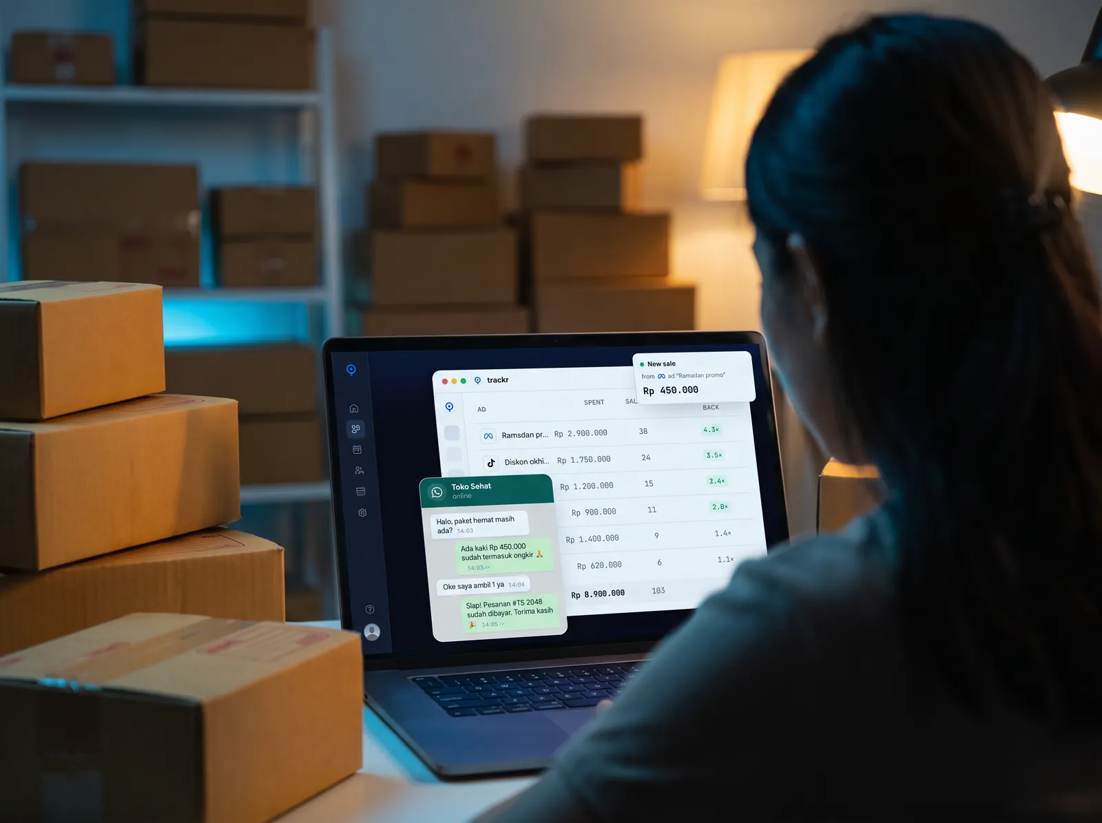 An online seller working late at night among packed parcels, checking orders on a laptop